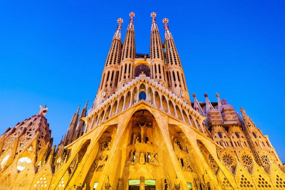 La imagen muestra la fachada frontal de la Basílica Expiatoria de la Sagrada Familia, un monumento icónico ubicado en Barcelona, España, y la obra maestra del arquitecto Antoni Gaudí. La fotografía se ha realizado al anochecer, con la fachada principal iluminada.