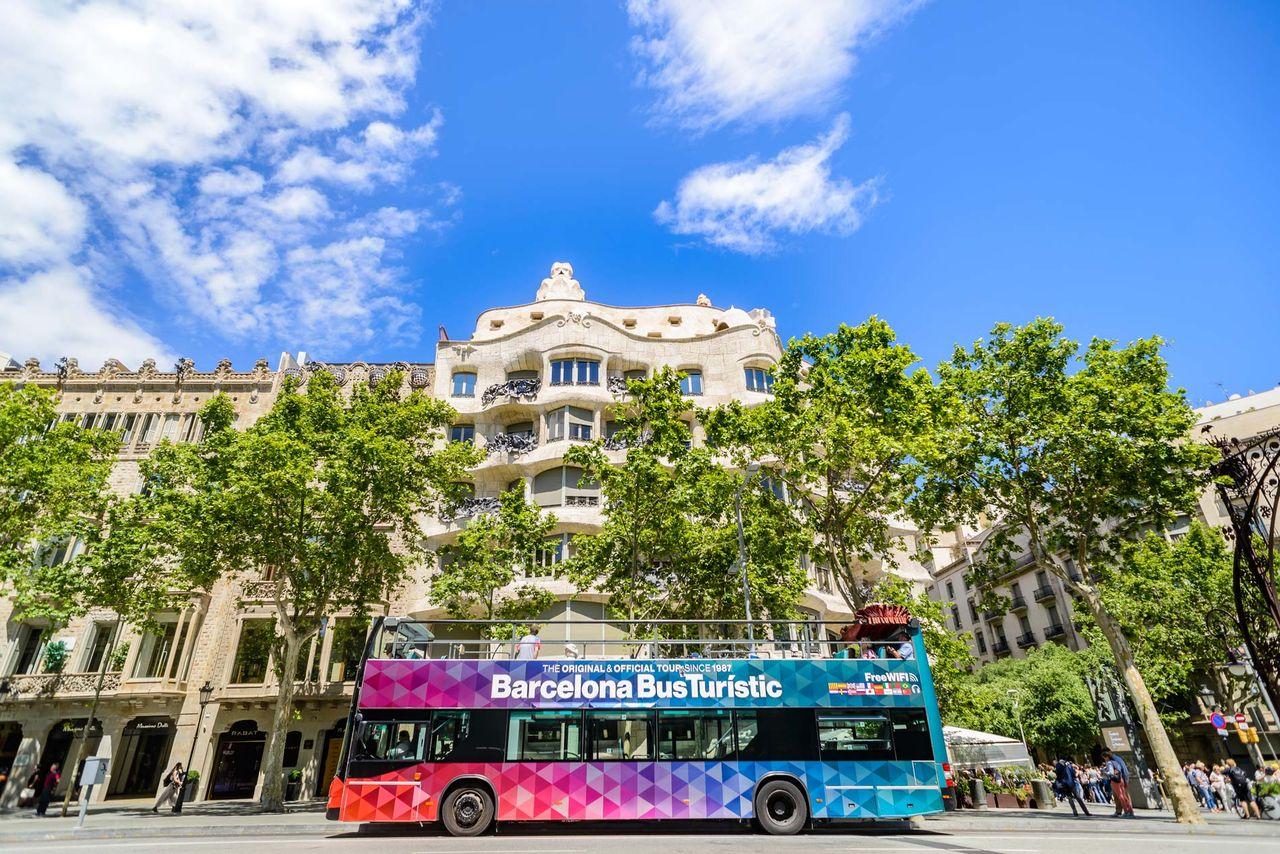 La imagen muestra el Barcelona Bus Turístic pasando justo por la fachada de La Pedrera. Este es el servicio oficial de autobús turístico de dos pisos (hop-on hop-off) de la ciudad.